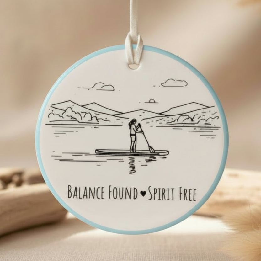 Circular pendant with a design of a person paddleboarding and text 'Balance Found Spirit Free' on a light blue background.